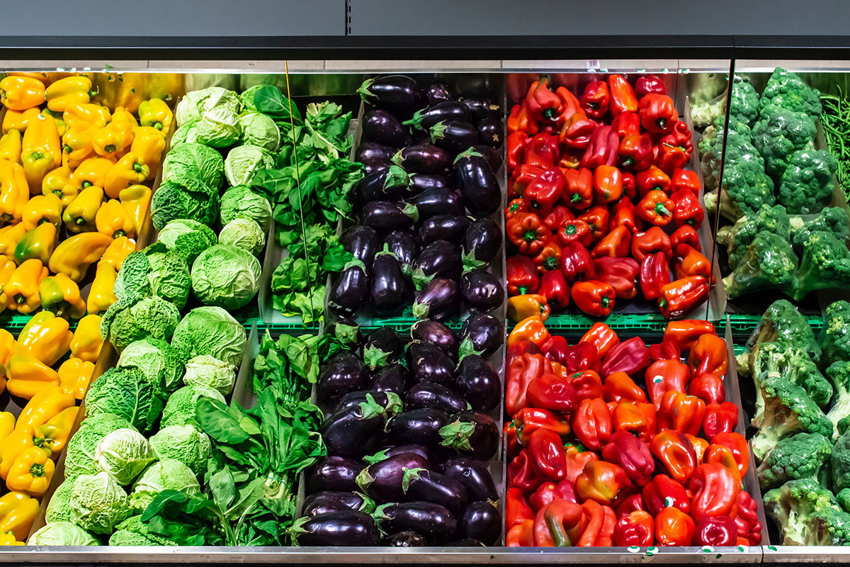 Grocery store produce section, with rows of yellow peppers, cabbage, eggplant, and broccoli.