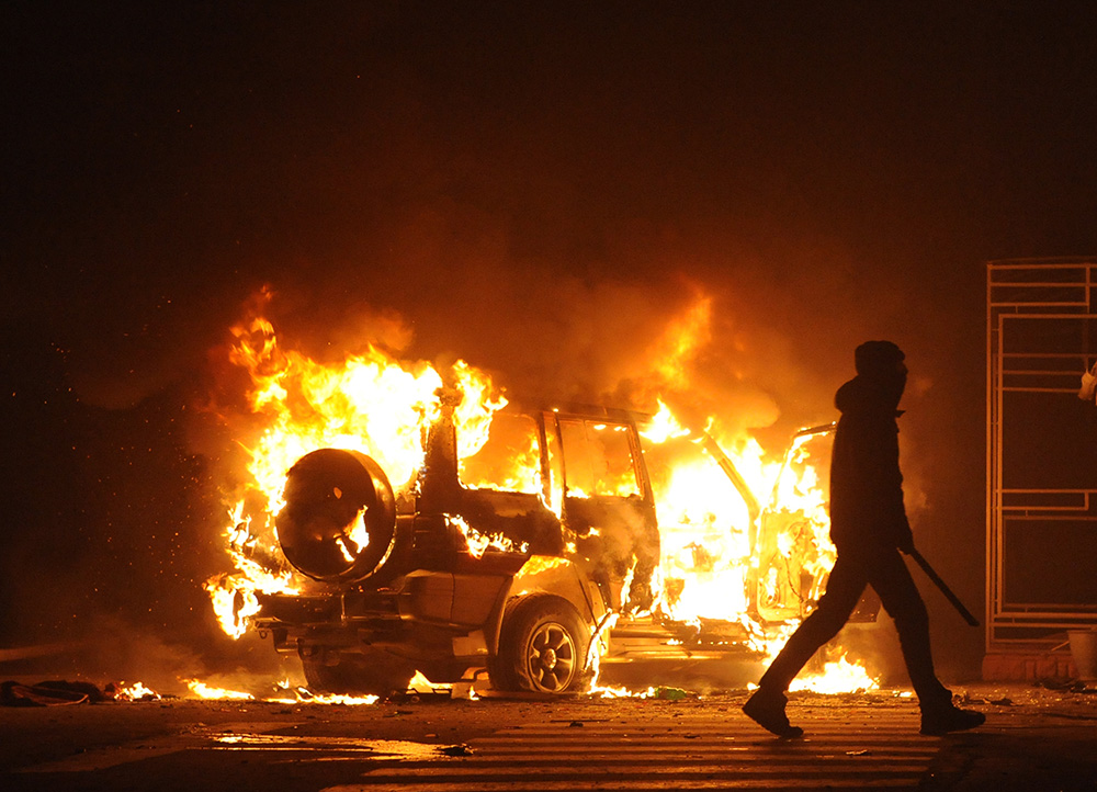 A silhouetted figure carrying a baton walks away from a burning car.