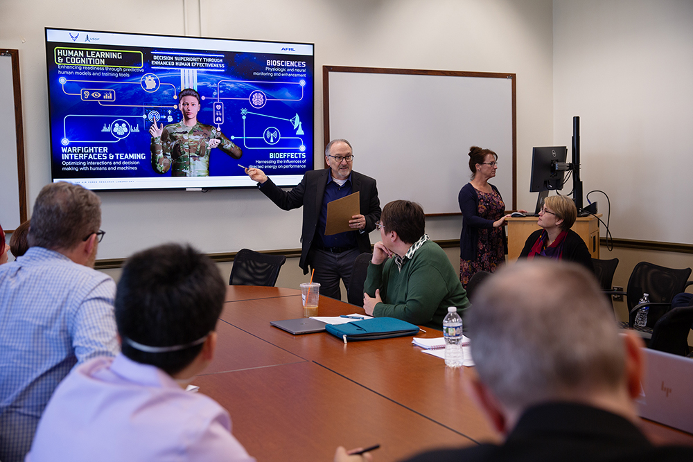 A man points to an image of an armed forces member on a projector screen in a full conference room.