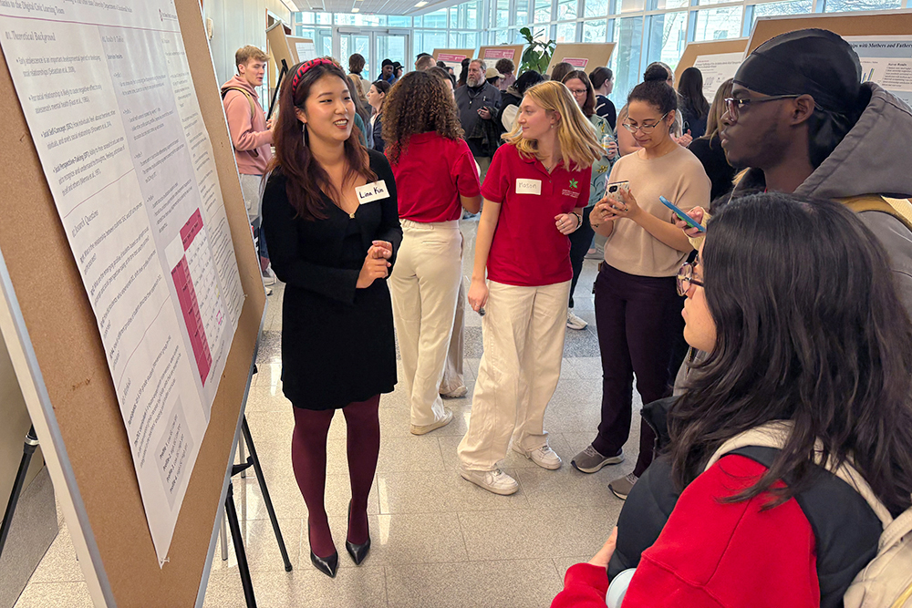 A student researcher speaks to a group of people gathered around her poster in a crowded building lobby.
