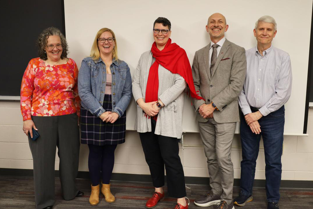 Five people stand smiling in front of a classroom blackboard