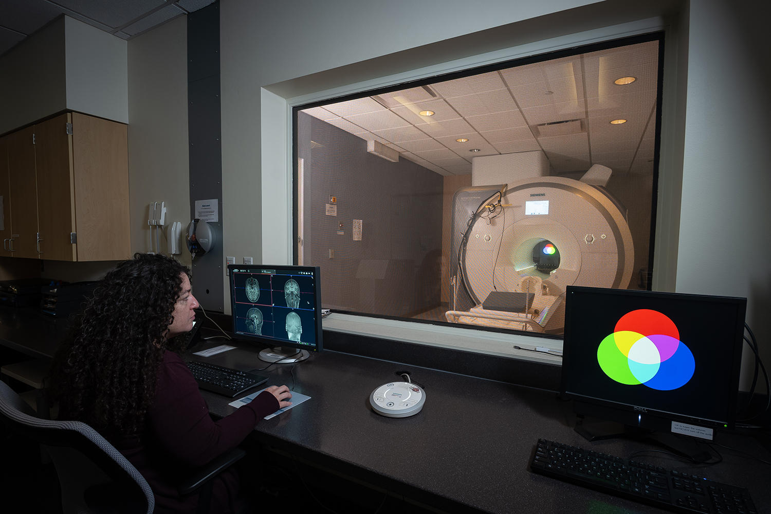 A woman watches a computer monitor with imagery of a human brain as an fMRI machine, visible through the window in front of her, operates.