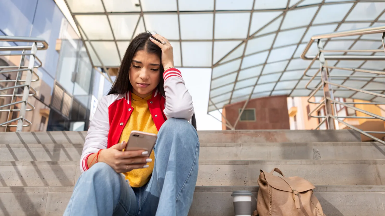 A young woman looks distressed as she sits on steps outside and looks at her smart phone.