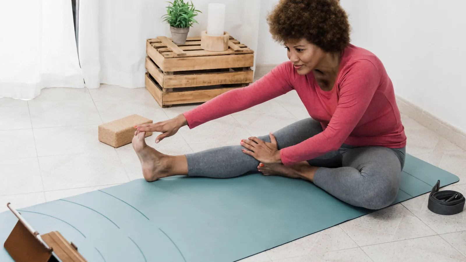 A woman stretches on a yoga mat while watching an instructor on her tablet.