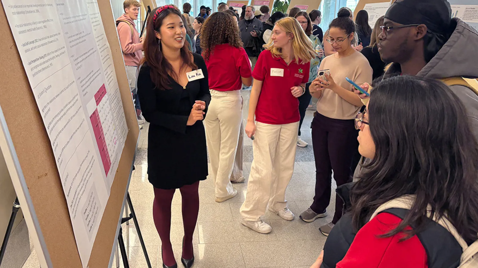 A student researcher speaks to a group of people gathered around her poster in a crowded building lobby.