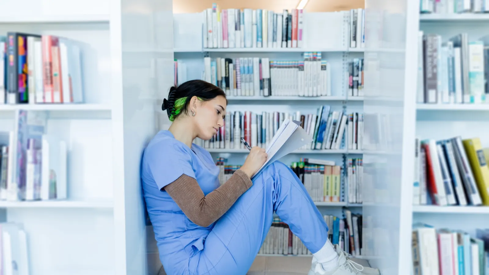 A woman in blue scrubs sits and reads a notebook between full bookshelves