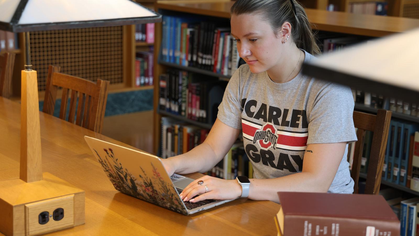 A student works on a laptop while surrounded by books in a library