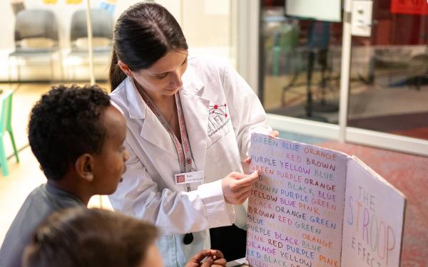A woman in a white lab coat holds up a placard with words written in multiple colors as a group of children looks on