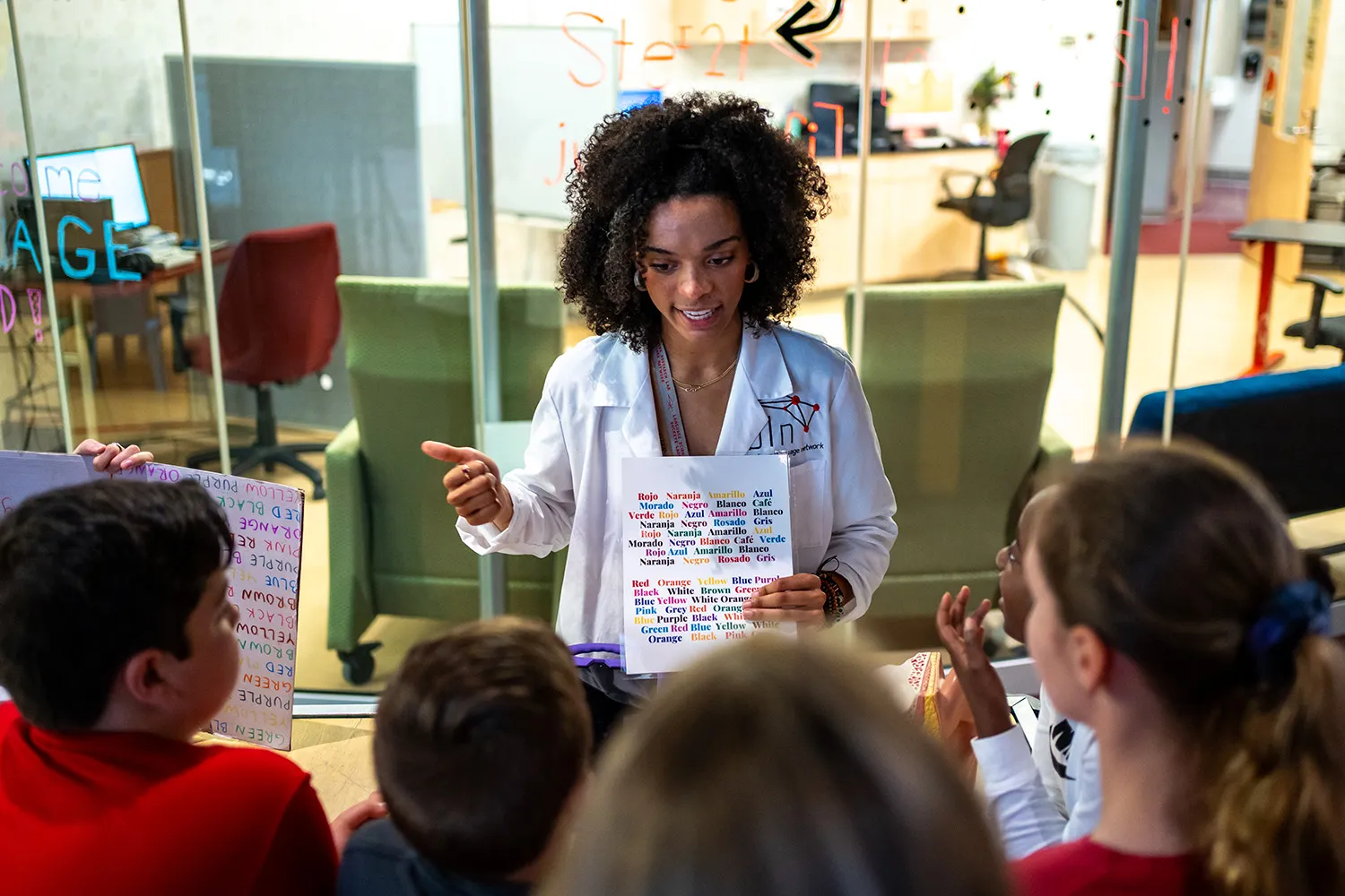 A woman in a white lab coat holds up a placard with words written in multiple colors as a group of children looks on
