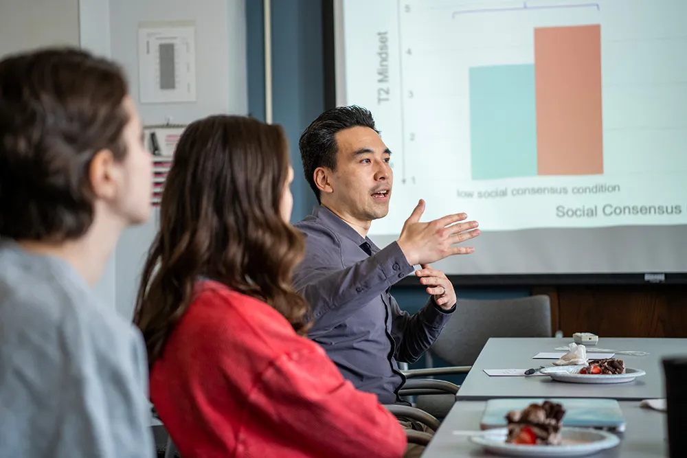 Kentaro Fujita speaks to students seated around a table while a graph is displayed on the screen behind him.