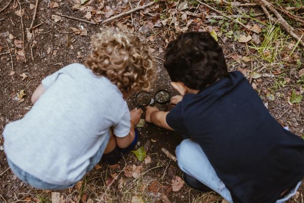 Two children use magnifying glasses to examine dirt on the ground.