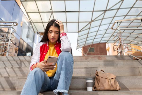 A young woman looks distressed as she sits on steps outside and looks at her smart phone.