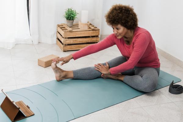 A woman stretches on a yoga mat while watching an instructor on her tablet.