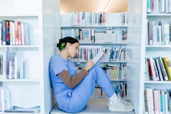 A woman in blue scrubs sits and reads a notebook between full bookshelves