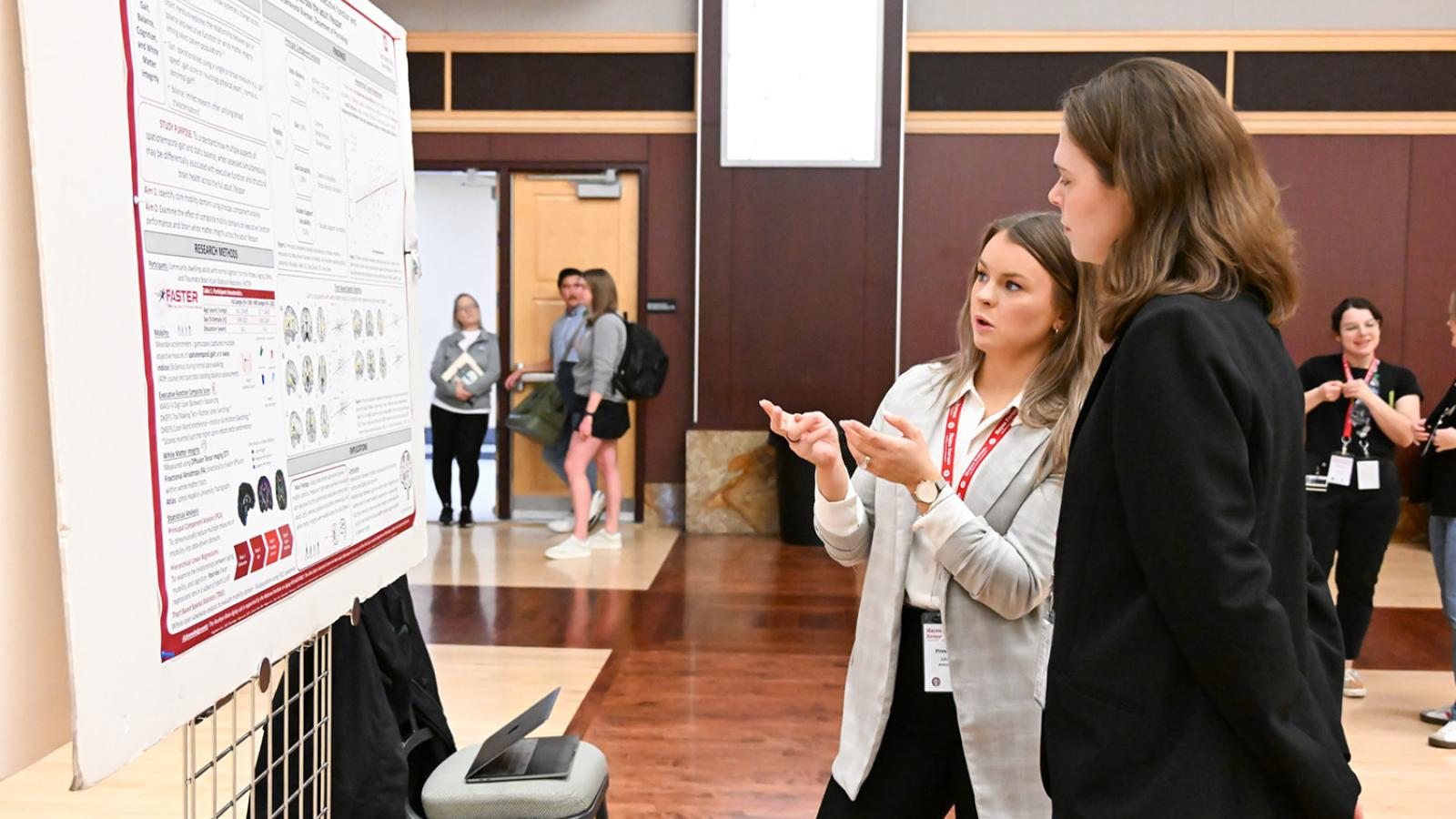A student presents a poster to a passer-by in an auditorium